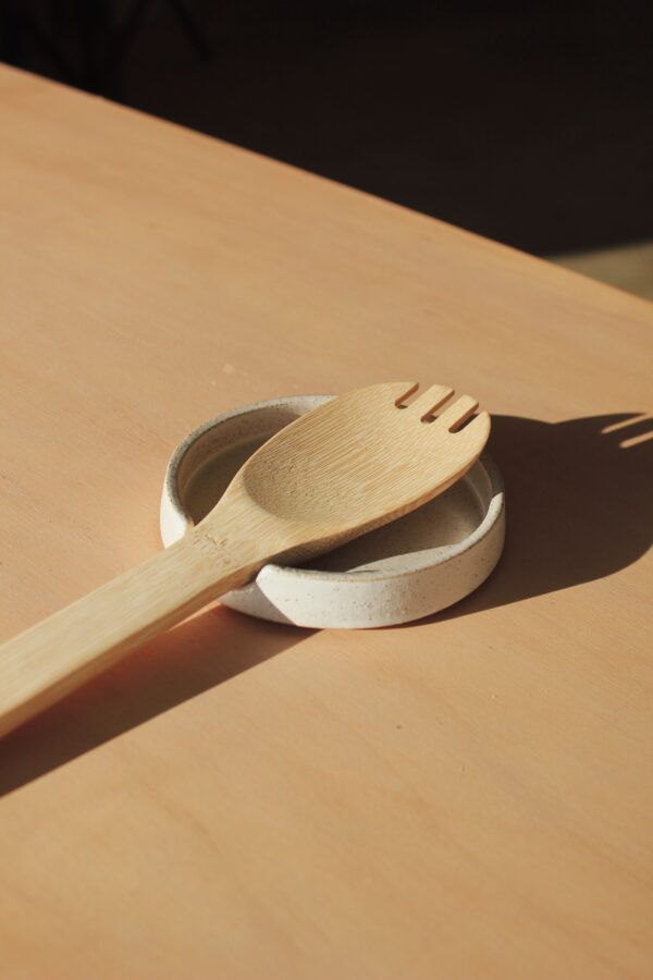 A white speckled spoon rest photographed against a maple wood background. The spoon rest features a wooden cooking spoon sitting in it.