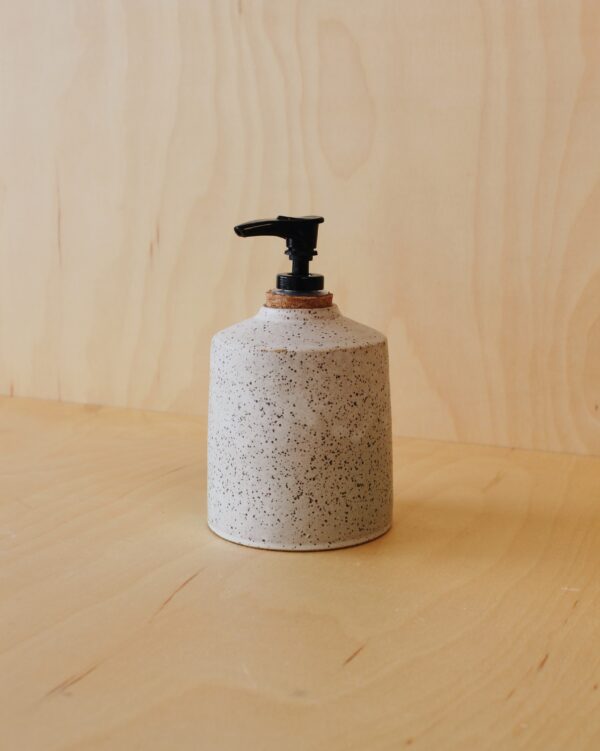 White speckled soap dispenser with a cork stopper pump. Photographed against a maple wood background.