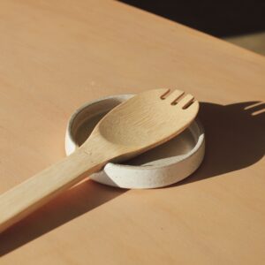 A white speckled spoon rest photographed against a maple wood background. The spoon rest features a wooden cooking spoon sitting in it.