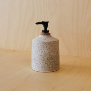 White speckled soap dispenser with a cork stopper pump. Photographed against a maple wood background.